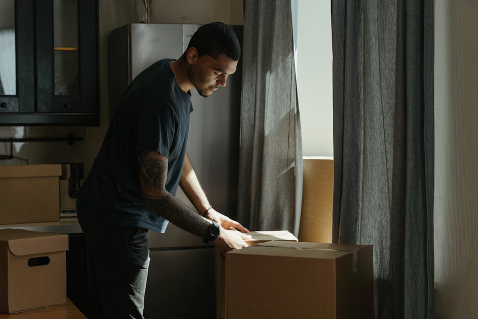 A man with a dark complexion, dressed in a black t-shirt and dark trousers, is inside a kitchen preparing for a home relocation. He is leaning over a large cardboard box placed on a wooden countertop, carefully unwrapping or inspecting packing materials. The kitchen has modern features with dark cabinets, a silver refrigerator in the background, and a window with partially drawn grey curtains allowing natural light to illuminate the area. The man is wearing a wristwatch, and his hair is short and neatly styled. To his left, there are additional cardboard boxes, some open and others sealed, indicating the packing process is underway. The scene captures a moment of packing and moving logistics, typical of professional furniture transport and house removals. As part of the moving process, [COMPANY_NAME], such as Man with Van Downe, might be involved in managing packing or loading tasks to facilitate a smooth transition for clients. The environment suggests organized packing, ensuring item protection during transport and proper planning for home relocation within or from Downe.