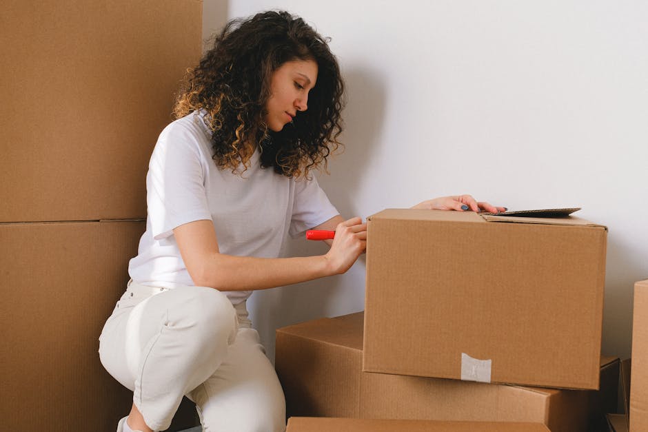 A woman with curly brown hair, dressed in a white t-shirt and light-coloured trousers, is kneeling on the floor inside a property, surrounded by several cardboard moving boxes in different sizes. She is using a red marker to label one of the boxes, which is placed on top of other packed boxes. The boxes are sealed with grey packing tape and appear to be prepared for a house relocation. Behind her, a plain white wall and a partially visible door frame suggest the interior of a home, where she is likely organizing or preparing items for removal. In the background, an open vehicle or loading area for furniture transport may be partially visible, indicating a loading process typical for professional removals. The scene conveys the packing stage of house removals, with an emphasis on careful labeling and organized packing materials, in a setting consistent with services offered by Man with Van Downe.