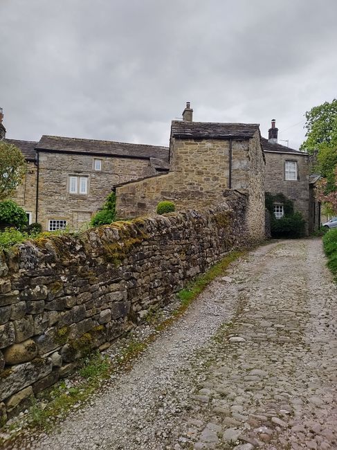 The image shows a historic stone cottage situated along a narrow residential street, with a weathered stone exterior and a steeply pitched slate roof featuring multiple dormer windows. The facade incl