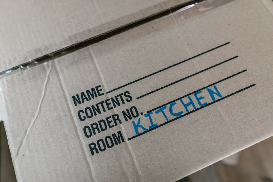 Close-up of a cardboard moving box labeled with black printed sections for 'Name,' 'Contents,' 'Order No.,' and 'Room,' with the word 'Kitchen' handwritten in blue marker in the 'Room' section. The box is part of a home relocation process, with other boxes possibly visible in the background. The surface of the box appears to be slightly textured, with clear tape sealing the top edges. The environment suggests an interior hallway or room during packing or moving preparations, with natural or ambient lighting illuminating the scene. This image illustrates the packing stage of furniture transport and house removals managed by Man with Van Downe, reflecting organized packing and moving logistics.