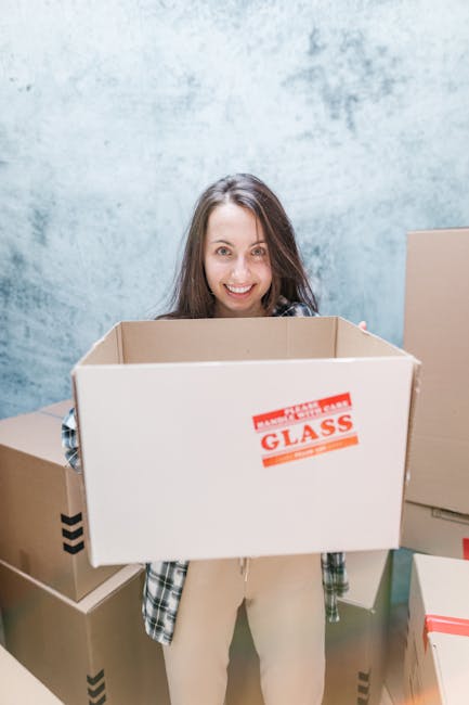 A young woman with long brown hair and a smile is standing indoors in front of a textured blue wall, holding an empty cardboard moving box with a red 'FRAGILE GLASS' sticker. She is dressed casually in a plaid shirt and light-colored trousers. Around her, there are several other packed moving boxes, some sealed with packing tape, positioned on the floor and stacked behind her. The scene appears to be part of a home relocation or packing process, with controlled lighting highlighting the boxes and the woman's face. This image reflects packing and moving activities typical of professional removals services like Man with Van Downe, emphasizing the careful handling of fragile items during furniture transport and home relocation procedures.