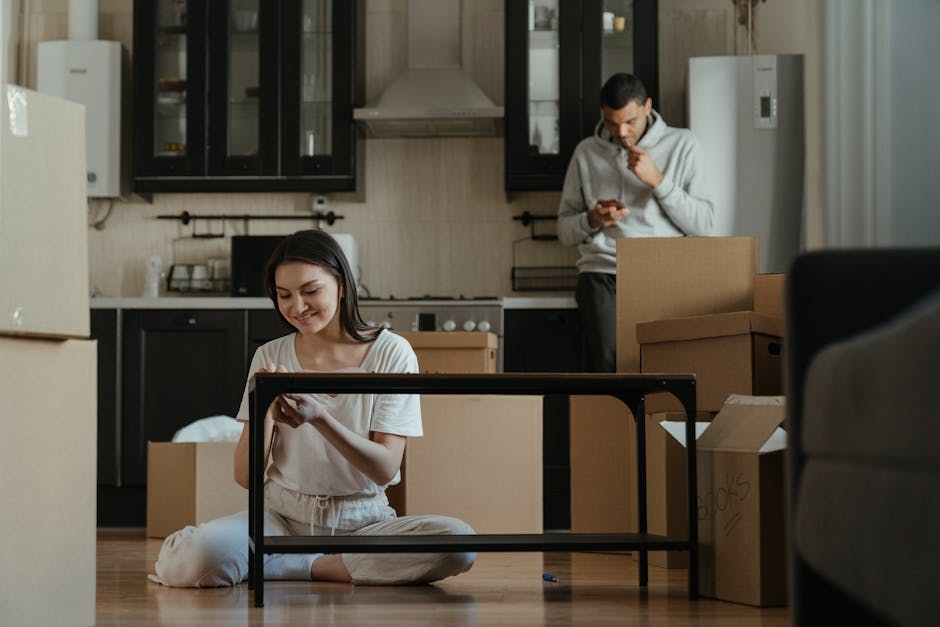 A young woman with dark hair sitting on the wooden floor of an interior room, smiling and looking at her phone, surrounded by open cardboard boxes, some filled with household items, near a black metal table frame. Behind her, a man wearing a light grey hoodie stands amidst more unpacked boxes, holding a phone to his ear, in a kitchen area with black cabinets, a stainless steel extractor hood, and a light-colored wall. The scene appears to be in the midst of a home relocation or packing process, with furniture and packing materials visible, supported by the professional removals services of Man with Van Downe.