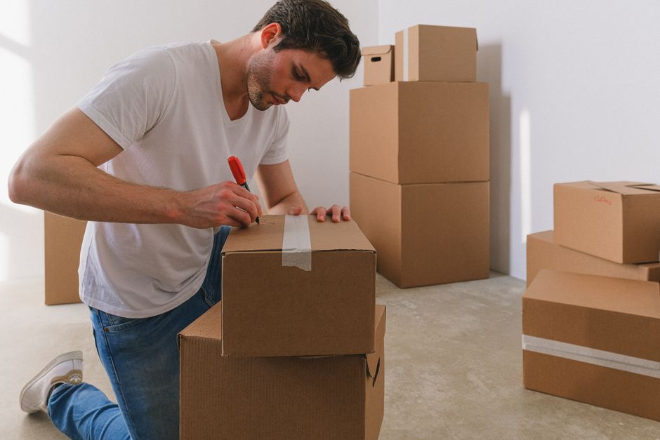 A young woman with dark hair sitting on the wooden floor of an interior room, smiling and looking at her phone, surrounded by open cardboard boxes, some filled with household items, near a black metal table frame. Behind her, a man wearing a light grey hoodie stands amidst more unpacked boxes, holding a phone to his ear, in a kitchen area with black cabinets, a stainless steel extractor hood, and a light-colored wall. The scene appears to be in the midst of a home relocation or packing process, with furniture and packing materials visible, supported by the professional removals services of Man with Van Downe.