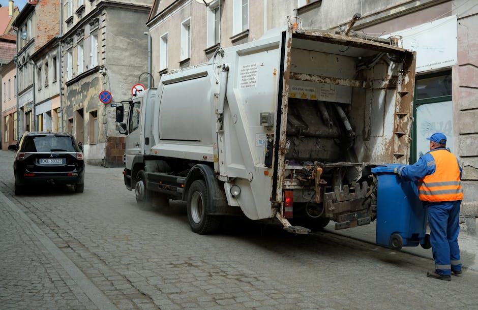 A street scene showing a worker from Man with Van Downe, dressed in a blue uniform with an orange high visibility vest and a blue cap, standing on the pavement beside a blue recycling bin while loading waste materials into a large white refuse truck with rusted and worn metal components at the rear. The truck is parked parallel to an old stone building with multiple windows, some of which are boarded up or have signs. Nearby, a black car is parked on the cobblestone street, and traffic signs are visible on the building wall. The scene is set during daytime with natural lighting, illustrating the process of waste collection as part of house removal or bulky waste disposal services, indicating a professional home relocation or clearance operation by [COMPANY_NAME].
