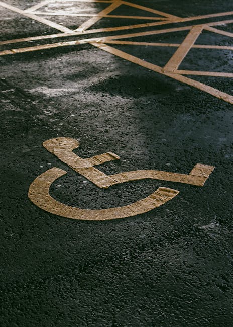 A close-up view of a wheelchair accessibility symbol painted in yellow on a dark, slightly wet asphalt surface, with visible texture and reflections. In the background, there are painted stripes and lines indicating parking bay boundaries, partially obscured by shadows. The scene suggests an outdoor parking area, possibly adjacent to a residential or commercial property awaiting or accommodating home relocation or furniture transport activities. The image highlights the designated accessible parking space, which is relevant for planning house removals or moving services by Man with Van Downe, especially when considering parking and access logistics for a smooth loading process during a house move.
