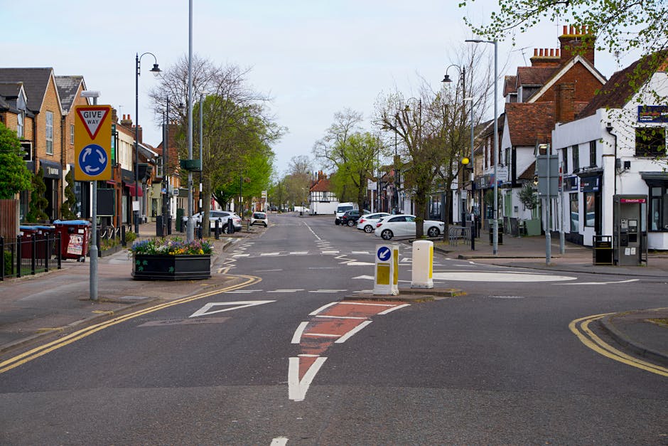 A quiet street in Downe village showing a well-maintained road with clear road markings, including a pedestrian crossing and a central traffic island with two yellow and white bollards. On the left side, there is a row of residential and small commercial buildings with traditional brick facades, some with windows and doors, and a few with shopfronts. Several cars are parked along both sides of the street, and a small vehicle is visible in the distance. Trees with budding leaves line the sidewalks, indicating springtime. A street lamp and a Give Way traffic sign with additional roundabout guidance are positioned near the central island. The scene appears to be during daytime with overcast sky, providing natural, diffused lighting. This setting illustrates an accessible, calm environment relevant to house removals, with potential for loading and unloading of furniture and moving boxes in a typical British village context, as managed and observed by Man with Van Downe during the home relocation process.